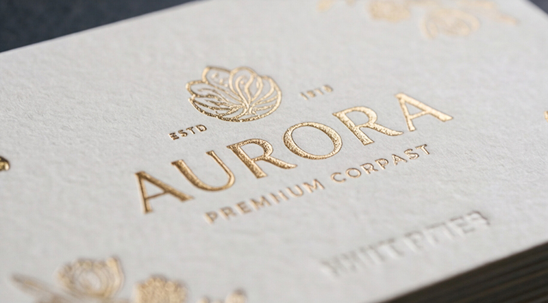 Close-up of a white textured card with gold foil embossing: a decorative emblem above the word 'AURORA' and 'PREMIUM' text below.