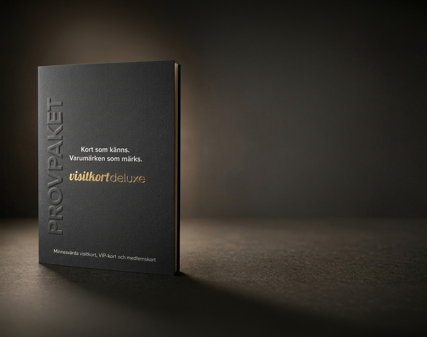 Dark matte booklet standing on a table with embossed vertical text on the left and gold 'visitkort deluxe' branding, in Swedish.